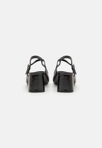 Black patent leather slingback heels with a chunky square heel and adjustable buckle strap, featuring a smooth texture and minimalistic design.
