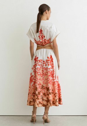 Femeie cu coadă de cal brunetă, purtând o rochie midi albă cu imprimeu floral roșu și o curea lată maro, stând în fața unui perete alb simplu.