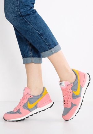 Personne portant un jean bleu retroussé et des baskets Nike rose, jaune et grises, debout avec un pied légèrement levé.