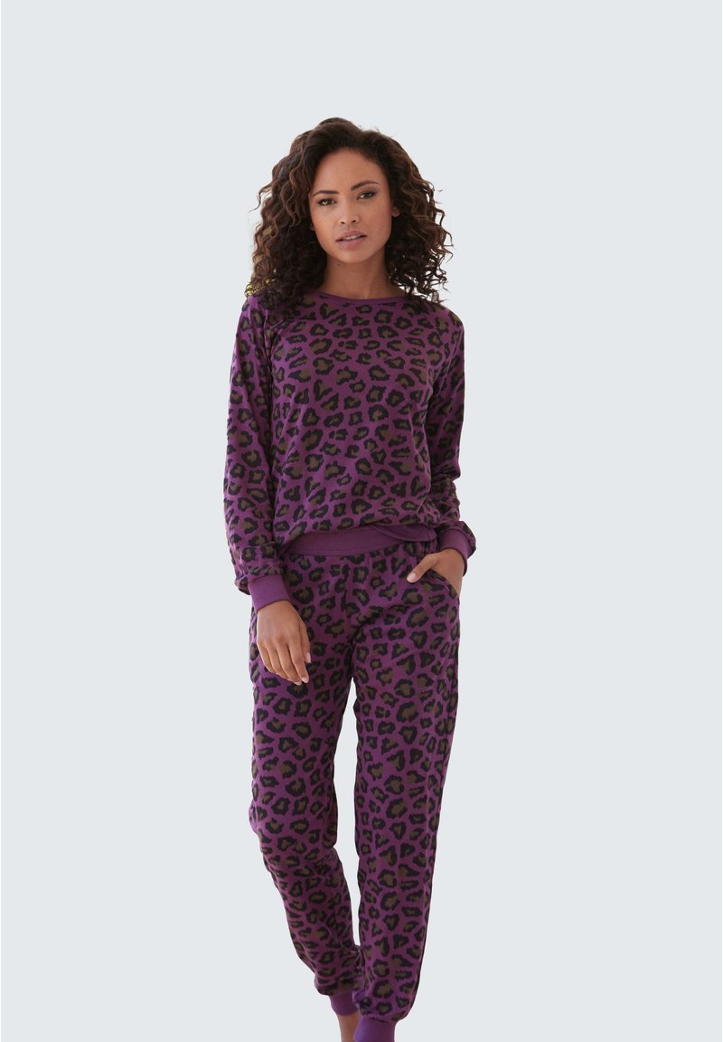 Donna con felpa e pantaloni jogger viola con fantasia leopardata coordinati, in piedi con una mano in tasca contro uno sfondo semplice.