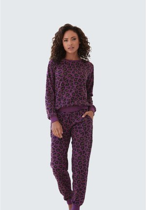 Donna con felpa e pantaloni jogger viola con fantasia leopardata coordinati, in piedi con una mano in tasca contro uno sfondo semplice.