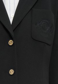 Blazer noir avec un emblème brodé sur la poche, des accents de boutons dorés et un tissu texturé. Présente un design classique et structuré.