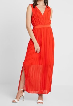 Femme portant une robe maxi rouge plissée sans manches avec une fente sur le côté et des sandales blanches à talons ouverts.