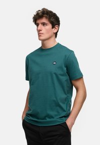 Tyrkise bomull t-skjorte med rund hals, korte ermer og en liten blå logo på brystet. Enkel design, myk tekstur.