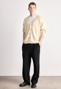 Bézs színű kötött kardigán mély V-nyakkivágással, hat nagy gombbal, bordázott mandzsettákkal és szegéllyel. Fekete, bő szabású nadrággal és fekete cipővel párosítva.