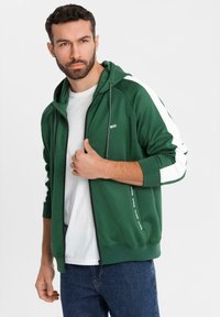 Grüne Zip-Hoodie mit weißen Seitenpaneelen; verfügt über eine Kapuze mit Kordelzug und weichem Stoff. Kombiniert mit einem weißen T-Shirt und blauen Jeans.