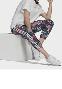 Blommiga leggings med mörk bakgrund, prydda med ett geometriskt mönster, vita ränder på sidorna, kombinerade med en lös vit t-shirt och vita sneakers.