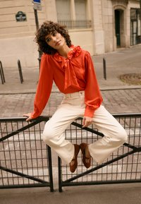 Blouse rouge vif avec col à volants et détail nœud papillon, associée à un pantalon écru taille haute et des ballerines marron. Fond uni.