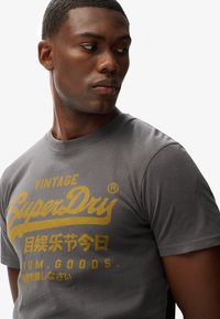 Graues Kurzarm-T-Shirt mit gelber grafischer Beschriftung "Vintage SuperDry" und mehreren Sprachen. Weicher Baumwollstoff, Rundhalsausschnitt.