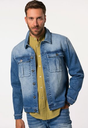 Denim-takkie i gradient blå nyanser, med to brystlommer, knappeåpning og en avslappet passform. Båret over en grønn skjorte.