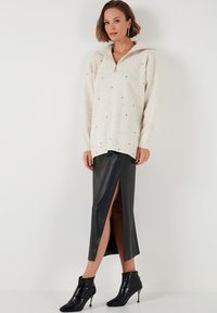Kremowy, oversize'owy sweter zapinany na zamek z srebrnymi akcentami; połączony z czarną, skórzaną spódnicą z wysokim rozcięciem i czarnymi botkami za kostkę.