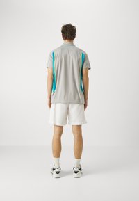 Šedé sportovní polo tričko s tyrkysovými a limetkově zelenými bočními pruhy, spárované s bílými šortkami. Model viděn zezadu.