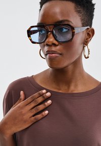 Przesadzone okulary przeciwsłoneczne w stylu żółwi z niebieskimi szkłami, w połączeniu z luźną brązową bluzką. Złote kolczyki w kształcie kół podkreślają wygląd.