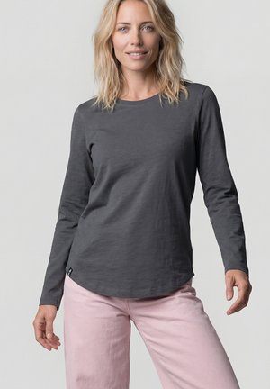 Frau mit schulterlangem blondem Haar, die ein dunkelgraues Langarmshirt und hellrosa Hose vor einem schlichten Hintergrund trägt.