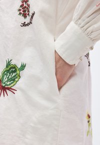 Λευκό πουκάμισο από ύφασμα με πολύχρωμα κεντημένα σχέδια λαχανικών και πλαϊνή τσέπη. Διαθέτει πιετέ μανίκια και διακοσμητικά κουμπιά στα μανίκια.