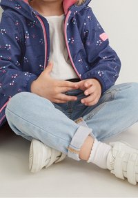 Veste à capuche bleu marine avec motifs floraux colorés, doublure rose et fermeture éclair à l'avant. Jean bleu clair et baskets blanches à scratch.