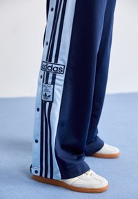 Mörkblå och ljusblå Adidas-träningsbyxor med tre ränder och märkespatchar, kombinerade med off-white sneakers på blått golv.
