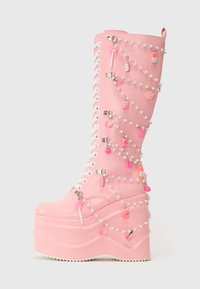 Bottes à plateforme roses avec une finition brillante, décorées de lacets blancs et de charmes sur des chaînes en perles, incluant des friandises et des papillons.