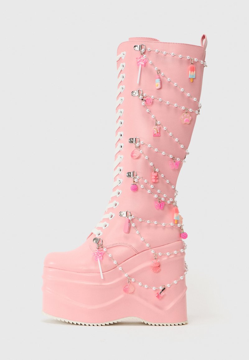 Bottes à plateforme roses avec une finition brillante, décorées de lacets blancs et de charmes sur des chaînes en perles, incluant des friandises et des papillons.