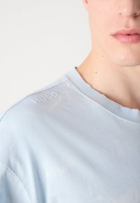 Vaaleansininen puuvillapaita, jossa on pyöreä kaula-aukko, hienovarainen haalistumiskuvio ja brodeerattu "HUGO" -logo vasemmassa olkapäässä.