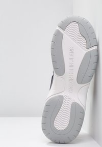 Bílá a šedá gumová podrážka tenisky Calvin Klein Jeans nakloněná proti bílé rohu.