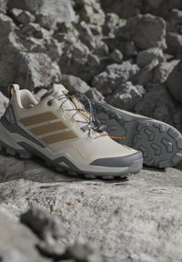 Scarpe da trekking beige chiaro e grigie con suole robuste, posizionate su un terreno roccioso, evidenziando il design pronto per l'outdoor e i sentieri.