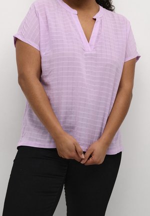 Personne portant un haut texturé violet clair à manches courtes et un pantalon noir, debout les mains légèrement jointes devant.