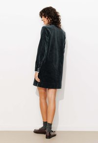 Robe en velours côtelé vert foncé à manches longues, coupe droite et détails de boutons aux poignets. Assortie avec des chaussures marron et des chaussettes grises.