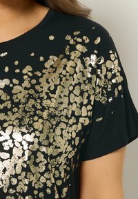 Sort T-shirt med et spredt metallic guld mønster. Designet inkluderer forskellige former med en struktureret finish og korte ærmer.