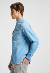 Camisa de mezclilla azul claro con mangas largas, dos bolsillos frontales, cuello con botones y dobladillo curvado. La tela tiene un acabado suave y ligeramente texturizado.