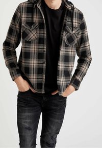Rutet flannelskjorte i sort, beige og creme; har to brystlommer og en hætte; iført over en sort t-shirt og slidte jeans.