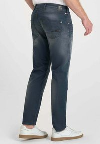 Jeans de mezclilla azul oscuro con corte ajustado, que presentan bolsillos traseros, un sutil desgaste y un parche con el logo en la cintura. Combinados con zapatillas blancas.