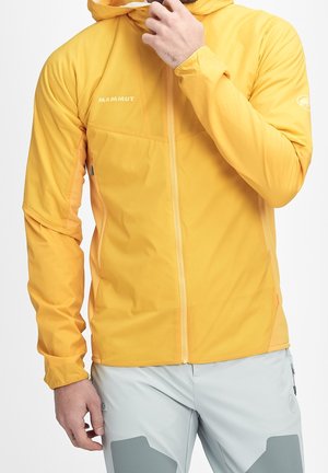 Hombre con chaqueta amarilla con capucha Mammut y pantalones gris claro, ajustando el cuello de la chaqueta con la mano derecha.