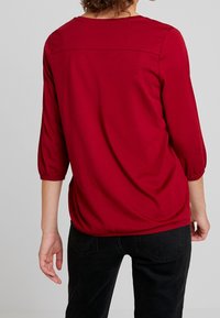 Kvinde iført en rød 3/4 ærmer top og sorte jeans, vist fra ryggen mod en ensfarvet baggrund.
