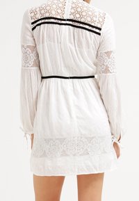 Biała koronkowa sukienka z długimi rękawami, zebranym w niewielkim pasem i czarnymi akcentami na ramionach. Design zawiera kwieciste wzory oraz detale z wiązaniami.