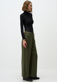 Maglietta nera a maniche lunghe con collo alto, abbinata a pantaloni larghi verde oliva e scarpe nere con finitura lucida.