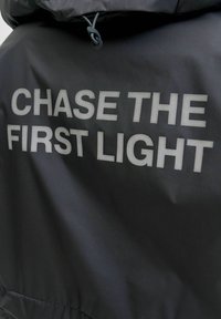 Czarna kurtka o błyszczącej teksturze z hasłem "CHASE THE FIRST LIGHT" dużymi, odbijającymi światło srebrnymi literami na plecach.