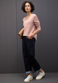 Maglione a V rosa chiaro, jeans scuri con risvolto e scarpe da ginnastica bianche. L'outfit presenta una texture liscia con un design semplice e casual.