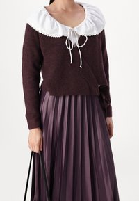 Kvinna iklädd en mörklila stickad omslagströja med en stor vit volangkrage, i kombination med en matchande plisserad kjol och håller i svarta väskremmar.