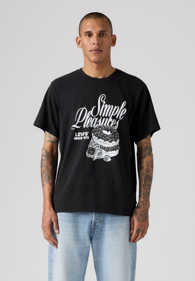 Άνδρας με τατουάζ φοράει μαύρο t-shirt Levi's με κείμενο "Simple Pleasures" και γραφικό με ντόνατ, σε συνδυασμό με ανοιχτό μπλε τζιν, στέκεται μπροστά σε γκρίζο φόντο.