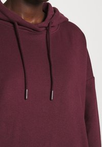Gros plan d'une personne portant un sweat à capuche bordeaux avec cordons, mettant en valeur la texture du tissu et la zone supérieure de la poitrine.