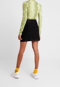 Osoba v zeleném tričku s hadím vzorem s dlouhým rukávem, černé sukni, bílých teniskách a žlutých ponožkách s černým textem, stojí na bílém pozadí.