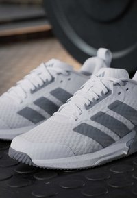 Baskets athlétiques blanches et grises avec trois bandes sur le côté, posées sur un sol noir de salle de sport près d'une plaque de poids.