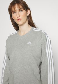 Šedá mikina s okrúhlym výstrihom, biele trojpruhy na rukávoch a vyšívané logo na hrudi. Mäkká textúra, voľný strih.