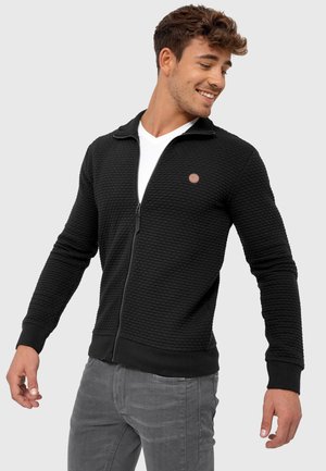 Schwarze Zip-Jacke mit strukturiertem Muster, mit hohem Kragen, gerippten Bündchen und einem kleinen braunen Logo-Patch auf der Brust.