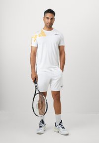 Witte sportshirt met gele geometrische patronen, gedragen met witte shorts. Het model houdt een tennisracket vast en draagt witte en blauwe sportschoenen.