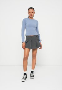 Młoda kobieta stoi w dopasowanej jasnoniebieskiej bluzie z długim rękawem, szarej plisowanej spódnicy z wiązaniem, białych skarpetach i czarnych sportowych butach za kostkę.