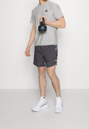 Hombre con camiseta gris de Adidas y pantalones cortos negros sosteniendo una pesa rusa negra, usando zapatillas blancas de Adidas sobre un fondo liso.