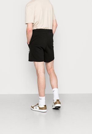 Hombre con pantalones cortos negros, camiseta beis, calcetines blancos de Nike y zapatillas Vans blancas y marrones, de pie con las manos en los bolsillos.