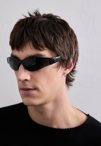 Lunettes de soleil noires au design élégant et enveloppant, avec une finition brillante. Elles comportent des détails de logo sur les branches et une monture légère.